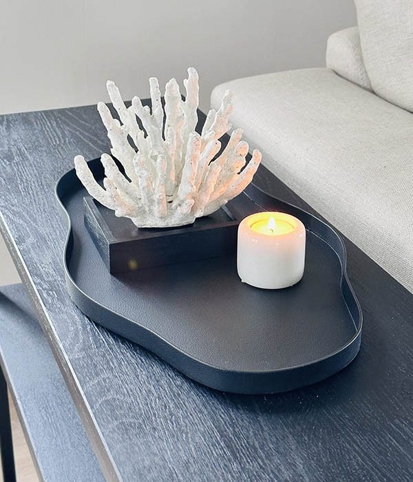 Zwart organisch gevormd dienblad met decoratief koraalornament en kaars op een zwarte eiken sidetable naast een beige bank