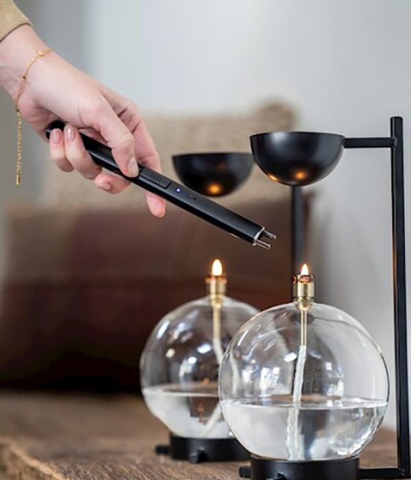 Hand met zwarte Peri Living oplaadbare aansteker boven twee glazen olielampen op een houten tafel